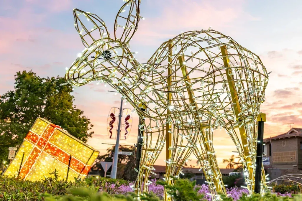 A large wireframe illuminated Christmas figure with a gift box in a civic garden bed at dusk